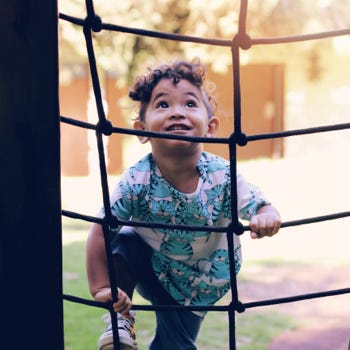 A toddler girl climbs up a climbing net at a playground.